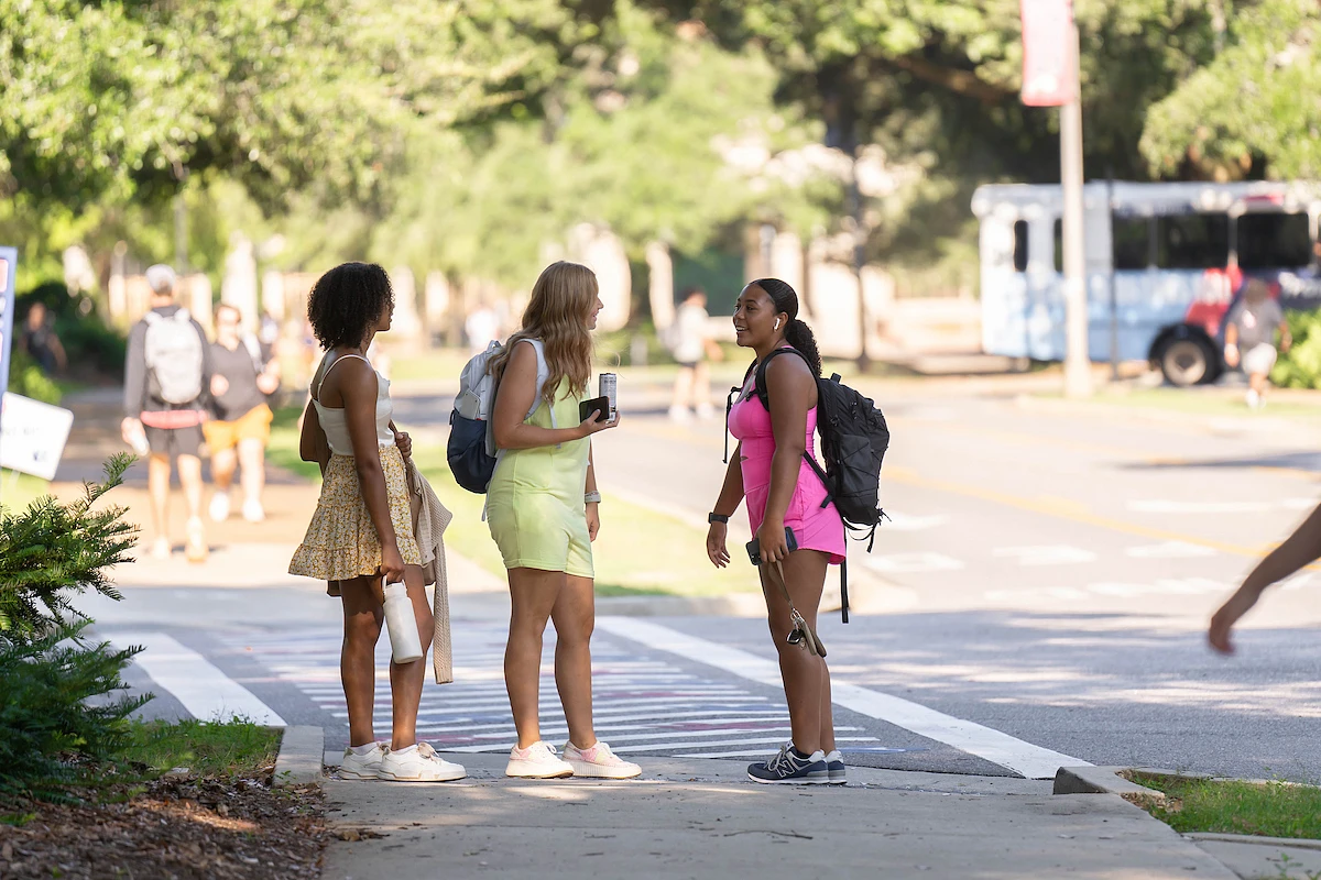 Three people with backpacks are chatting on a sunny sidewalk near a street.